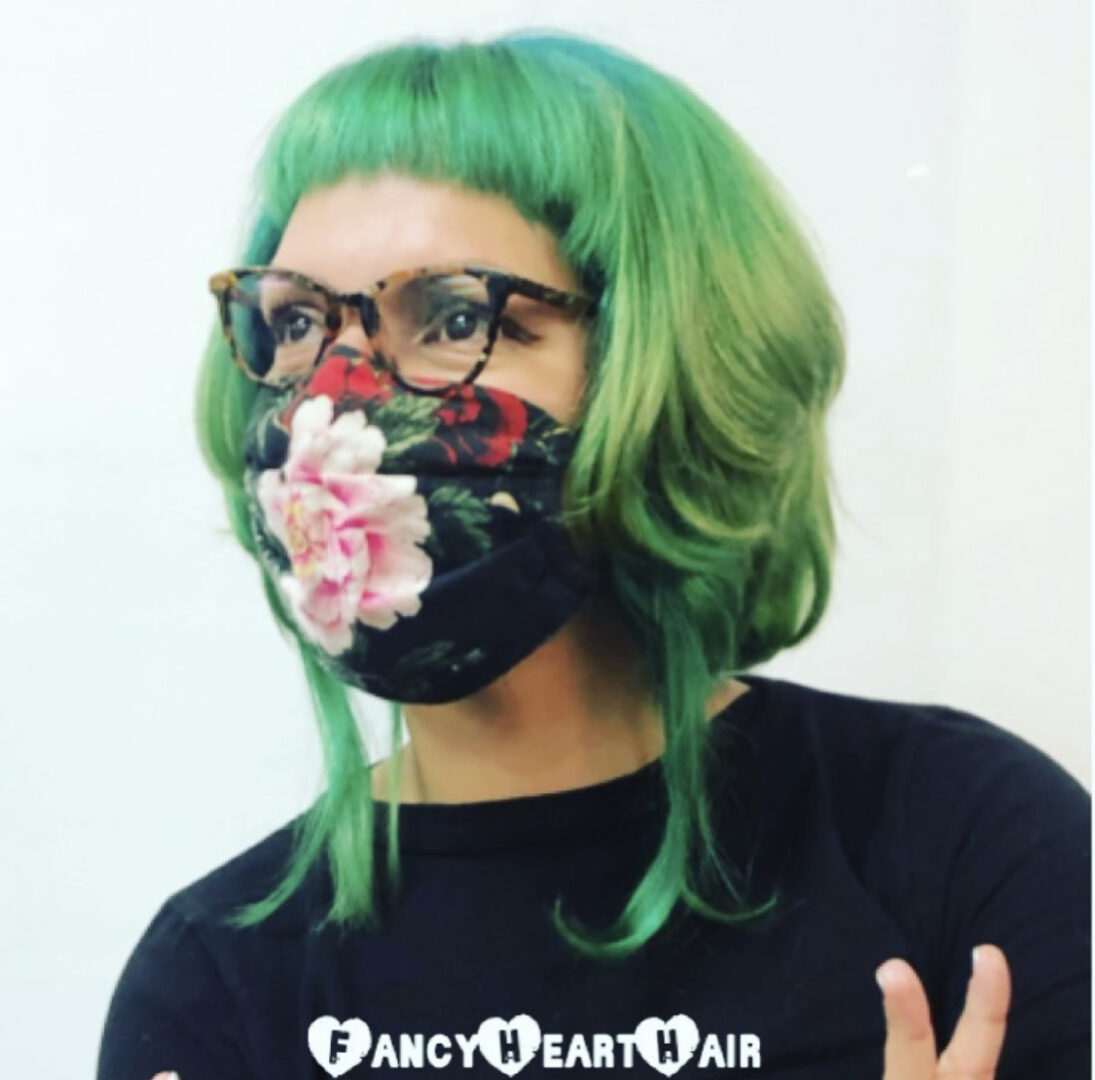 A person with green hair and glasses wearing a floral mask.